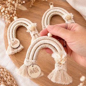 Könnte beinhalten: Ein Set aus drei Makramee-Regenbogen-Anhängern in Creme- und Goldtönen. Jeder Regenbogen besteht aus geflochtener Kordel, einer Holzperle, Spitzenbesatz und Trockenblumen. Ein rundes Holzschild mit der Gravur GLUCKS BRINGER ist jeweils befestigt.