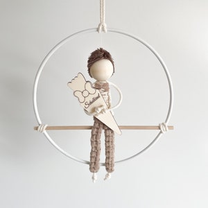 Op de afbeelding: Een macramé pop met een houten hoofd en armen, met een houten schooltuit met het woord "Schulkind". De pop hangt in een witte metalen hoepel met een houten deuvel en een wit touw.