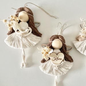 May include: Three handmade brown yarn angel ornaments with white yarn dresses and wings. The middle angel holds a wooden heart with the name "Klara" written on it.
