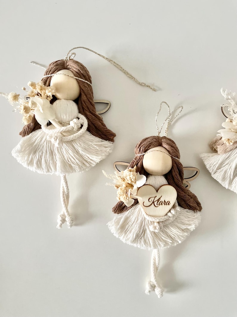 May include: Three handmade brown yarn angel ornaments with white yarn dresses and wings. The middle angel holds a wooden heart with the name "Klara" written on it.