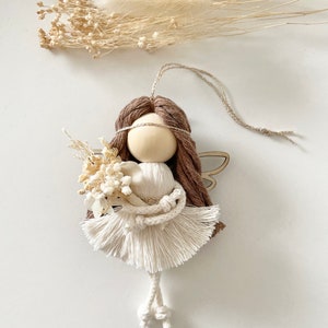 May include: A brown yarn angel doll with white yarn wings and a white yarn dress. The doll is holding a small bouquet of dried flowers.