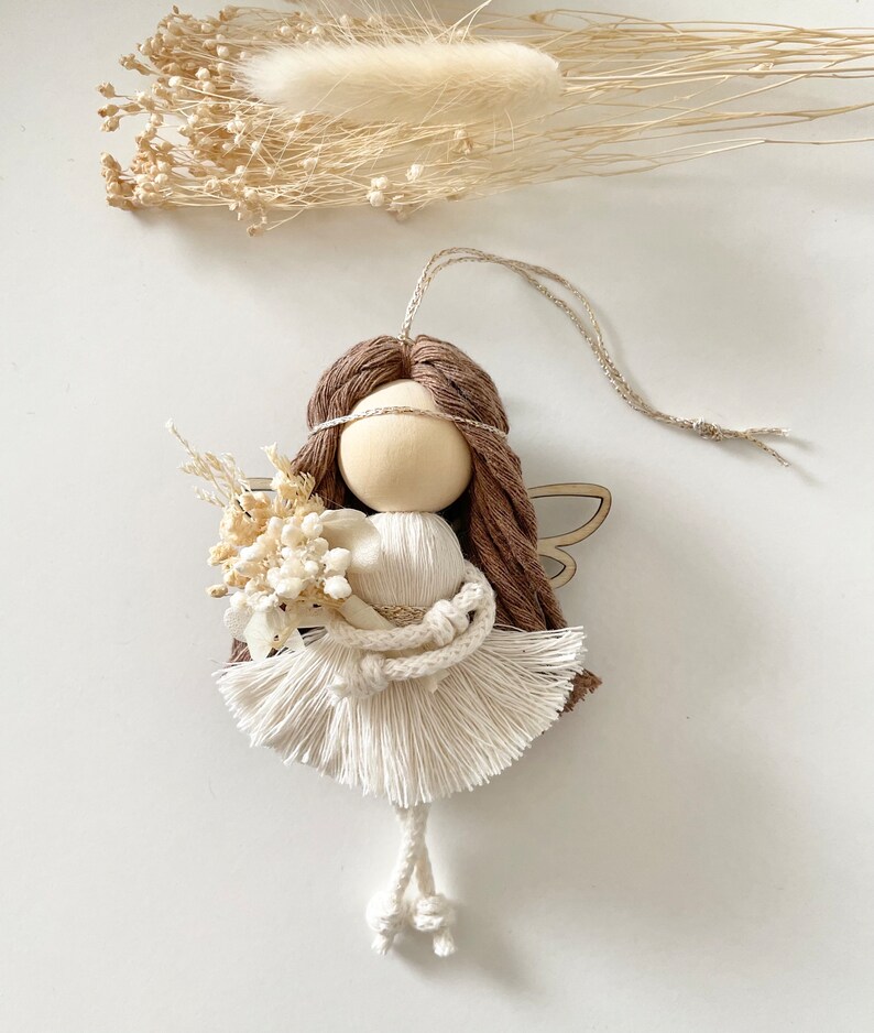 May include: A brown yarn angel doll with white yarn wings and a white yarn dress. The doll is holding a small bouquet of dried flowers.