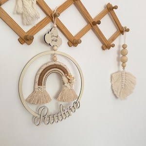 Könnte beinhalten: Ein runder Wandbehang aus Holz mit einem Makramee-Regenbogen in Beige- und Cremetönen, verziert mit Trockenblumen. Darüber hängt eine Holzwolke mit der Aufschrift "Du wirst geliebt", und unten ist der Name "Pauline" aus Holz ausgeschnitten.