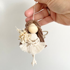 May include: A white yarn angel ornament with brown yarn hair, a wooden head, and a small bouquet of dried flowers. The angel has wings made of wood and is hanging from a string.