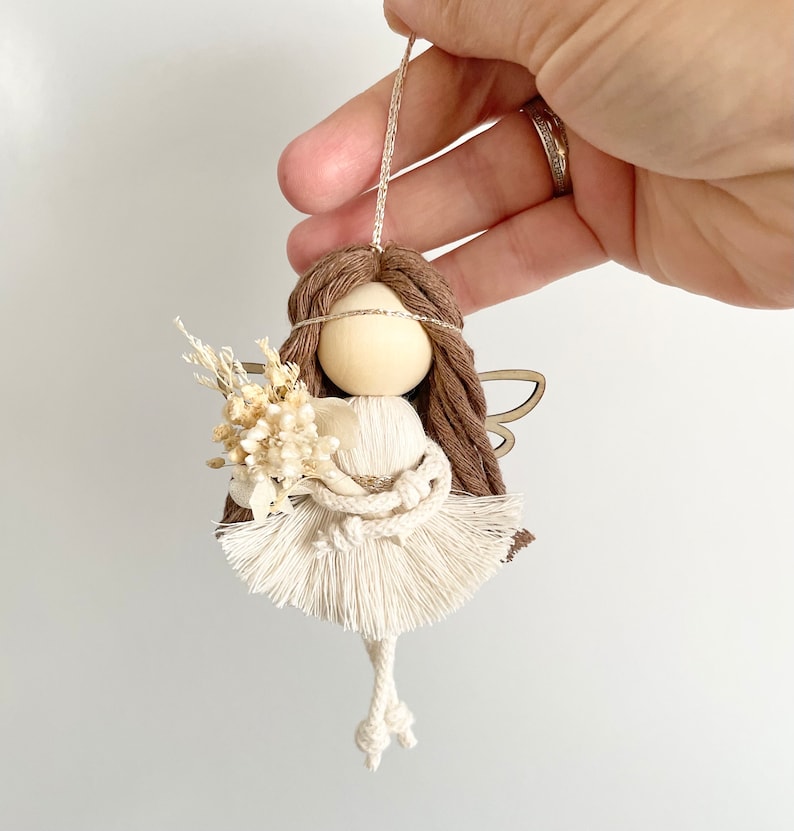 May include: A white yarn angel ornament with brown yarn hair, a wooden head, and a small bouquet of dried flowers. The angel has wings made of wood and is hanging from a string.