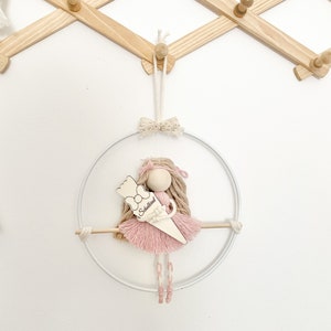 Op de afbeelding: Handgemaakte pop met een roze jurk en lang gevlochten haar, opgehangen in een witte metalen hoepel. De pop houdt een houten bord vast met de tekst "Schulkind". De pop hangt aan een houten wandrek.