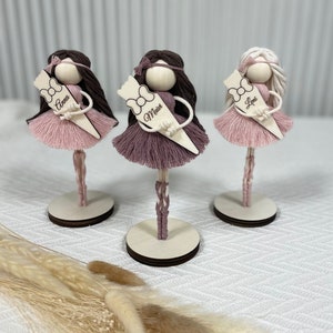 Op de afbeelding: Drie roze en witte garenpoppenfiguren met bruin haar en houten bases. Elke pop houdt een houten bordje vast met een naam: Anna, Mais en Leni.