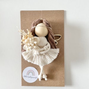 May include: A white and brown yarn doll with a white dress and brown hair. The doll is holding a bouquet of dried flowers. The doll is on a brown card with a white circle that says "littleplazaworld" with a rainbow inside.