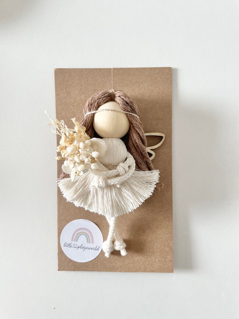 May include: A white and brown yarn doll with a white dress and brown hair. The doll is holding a bouquet of dried flowers. The doll is on a brown card with a white circle that says "littleplazaworld" with a rainbow inside.