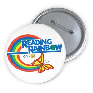 Puede incluir: Un botón blanco con el logotipo de "Reading Rainbow" en azul, rojo, amarillo y verde. Una mariposa colorida aparece debajo del texto. El botón tiene un respaldo de alfiler de metal.