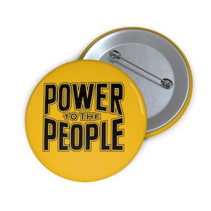 Può includere: Bottone giallo con la scritta nera "POWER TO THE PEOPLE". Il bottone è rotondo, lucido e ha una spilla argentata sul retro. Il testo è in grassetto e senza grazie.