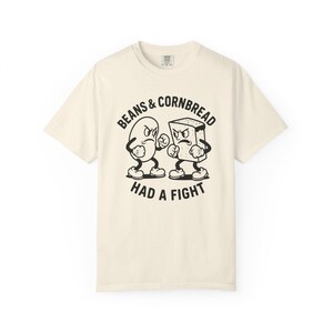 May include: Cream-colored t-shirt featuring a black and white cartoon illustration of a bean and a piece of cornbread engaged in a fist fight. The text "BEANS & CORNBREAD HAD A FIGHT" is printed above and below the characters.