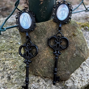 Könnte beinhalten: Ein Paar schwarze Ohrringe mit Schlüsseldesign. Jeder Ohrring hat einen ovalen, irisierenden Mittelstein, der in einen schwarzen Filigranrahmen gefasst ist. Der Schlüsselanhänger hängt darunter und erzeugt eine gotische Ästhetik.