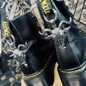 Könnte beinhalten: Ein Paar schwarze Lederstiefeletten mit strukturierter Oberfläche, gelben Rahmennähten und schwarzen Fersenschlaufen mit gelbem AirWair-Schriftzug. Jeder Stiefel ist mit einem silberfarbenen Ziegenkopf-Anhänger an der Fersenschlaufe verziert.