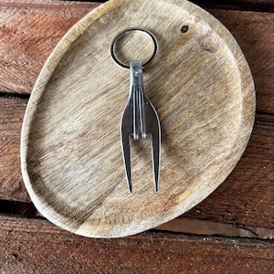 Pode incluir: Um porta-chaves de garfo de prata feito à mão com um design de dentes divididos. O porta-chaves é preso a um anel de prata. O garfo está sobre um prato oval de madeira com um padrão natural de grão de madeira.