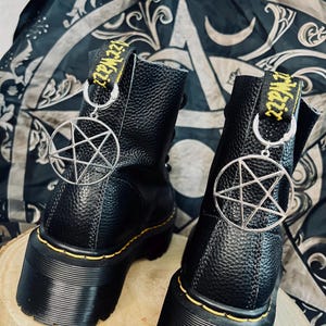 Könnte beinhalten: Ein Paar schwarze Plateau-Stiefel aus genarbtem Leder mit gelben Nähten entlang der Sohle und einer gelben Fersenschlaufe mit schwarzem Text. Jeder Stiefel ist mit einem silberfarbenen Pentagramm-Anhänger aus Metall an der Ferse verziert.