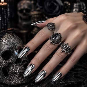 Könnte beinhalten: Drei silberfarbene Metallringe mit filigranen Verzierungen, einem zentralen ovalen schwarzen Stein und einem Totenkopfmotiv. Die Ringe werden an Fingern mit langen, spiegelnden silbernen Nägeln getragen und zeigen ein detailliertes Design.