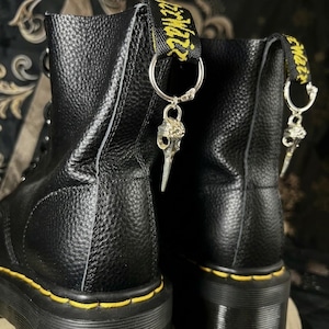 Könnte beinhalten: Ein Paar schwarze Lederstiefel mit strukturierter Oberfläche und gelben Nähten entlang der dicken Sohlen. Jeder Stiefel hat eine schwarze Fersenschlaufe mit gelbem Text und einen silberfarbenen Metallanhänger in Form eines Vogelschädels.