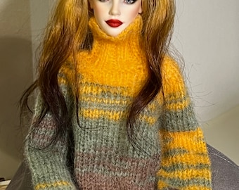 Può includere: Una bambola che indossa un maglione peloso a righe in tonalità di marrone, verde e giallo. Il maglione ha un collo alto e maniche lunghe.
