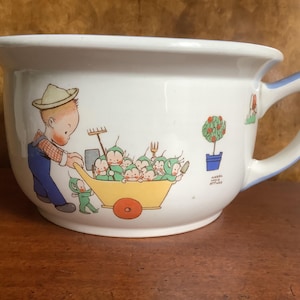 May include: A white ceramic potty with a blue rim and handle. The potty features a cartoon illustration of a boy in overalls pushing a wheelbarrow full of little green people. The text "Made in England" is visible on the side.