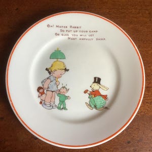 May include: A vintage white plate with a red rim, decorated with a charming illustration. The image shows a girl with an umbrella, a fairy, a doll, and a rabbit in a top hat. The plate has the text: "Oh! Mister Rabbit, Do put up your gamp, Or else you will get, Most awfully damp."