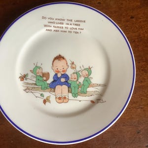 May include: A vintage white plate with a blue rim. The plate has a charming illustration of a child and fairies. The text on the plate reads, "Do you know the laddie who lives in a tree with fairies to love him and ask him to tea?"