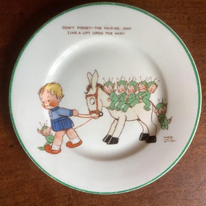 May include: A white porcelain plate with a green rim features a whimsical illustration of a child leading a donkey carrying fairies. The text on the plate reads "Don't forget-the fairies may like a lift upon the way!"