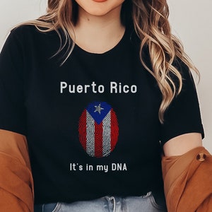 Puede incluir: Una camiseta negra con un gráfico blanco de la bandera puertorriqueña en un diseño de huella digital. El texto "Puerto Rico" y "It's in my DNA" está impreso en la camiseta.