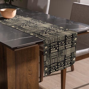 May include: A black and beige patterned table runner with a geometric design. The runner is draped over a dark table, showcasing a repeating pattern of lines, shapes, and symbols. The runner adds a decorative touch to the dining table.