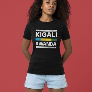 May include: A black t-shirt with white text that reads "KIGALI RWANDA LAND OF A THOUSAND HILLS" with the Rwandan flag colors in a stripe above the word "RWANDA".