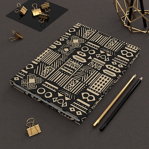 May include: A closed journal with a black cover featuring a tan geometric pattern. Gold and black pencils, along with gold binder clips, are arranged around the journal on a dark gray surface. The journal's design is reminiscent of tribal art.