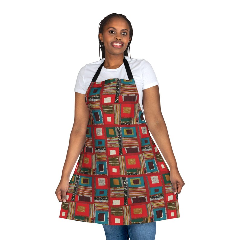 May include: An apron with a colorful geometric pattern. The apron features a red background with squares and rectangles in shades of teal, green, brown, and white. It has a black neck strap and is worn by a person.