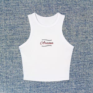 May include: White sleeveless crop top with the word "Arizona" in maroon script, stitched with baseball-style seams. The top is made of a soft, lightweight material, perfect for warm weather. The top is displayed on a blue denim background.
