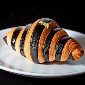 May include: A chocolate croissant with alternating dark and light brown stripes, topped with a gold leaf, rests on a white plate with blue stripes. The pastry is a delicious treat.