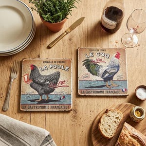 Könnte beinhalten: Zwei quadratische Untersetzer im Vintage-Stil mit Hahn- und Hühnerillustrationen. Die Untersetzer zeigen französischen Text und stehen auf einem Holztisch mit einer Weinflasche, einem Glas und Brot.