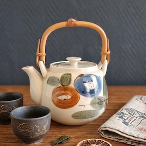 Puede incluir: Una tetera de cerámica color crema con asa curva de bambú y tapa, decorada con motivos de frutas pintados a mano en naranja y azul con hojas verdes. Dos tazas de cerámica gris oscuro con ilustraciones doradas de grullas y pinos.