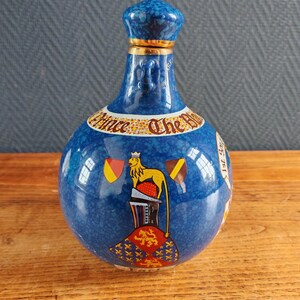 May include: A blue ceramic bottle with a textured surface and a matching stopper. The bottle features a gold band and a design with a lion, flags, and the text "Prince The Blue".