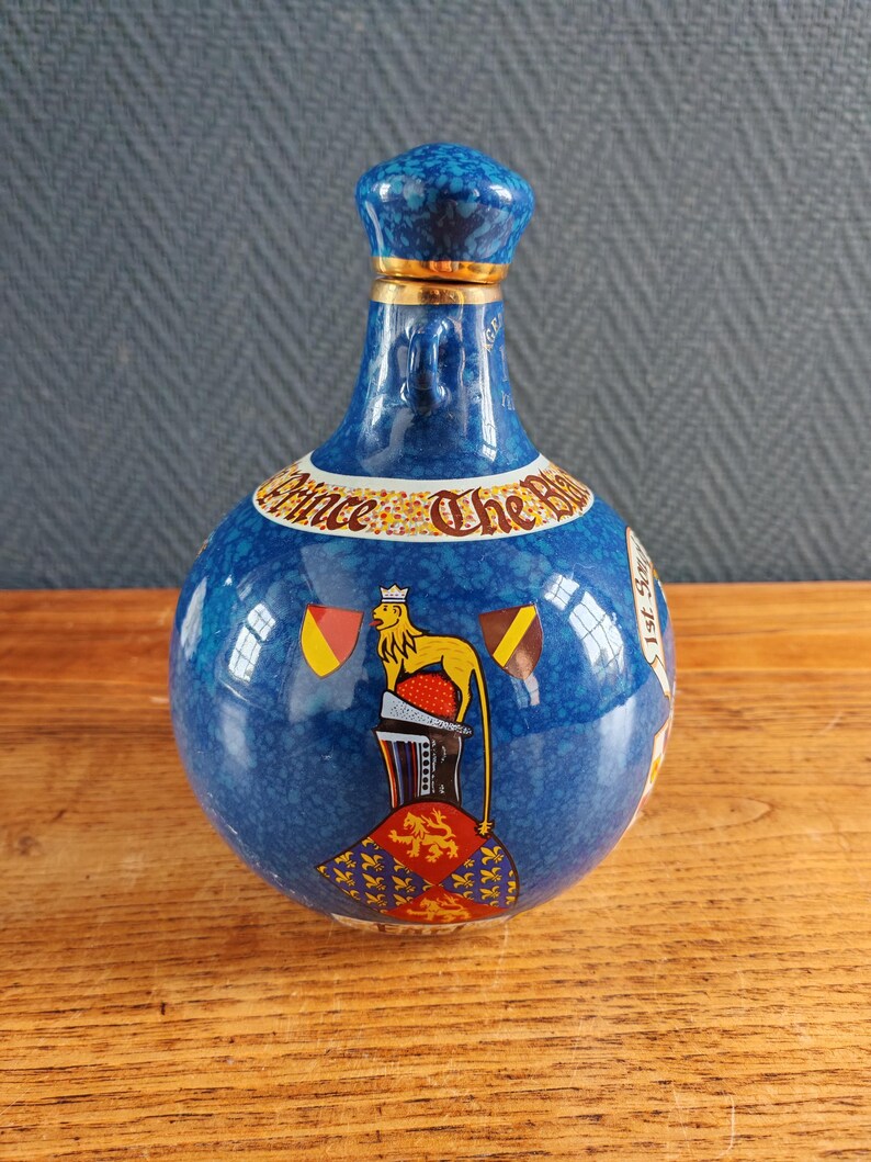 May include: A blue ceramic bottle with a textured surface and a matching stopper. The bottle features a gold band and a design with a lion, flags, and the text "Prince The Blue".