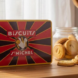 Op de afbeelding: Een rechthoekige blikken doos met St. Michel-koekjes met een rood en zwart ontwerp, een pot koekjes en een paar koekjes op een houten oppervlak. De doos bevat de woorden "BISCUITS" en "ST MICHEL" met een hanenembleem.