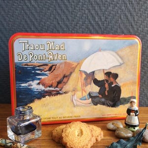 May include: A rectangular metal tin labelled Traou Mad De Pont-Aven featuring a coastal illustration, a glass inkwell, a fountain pen, two round biscuits, a small ceramic figurine in traditional attire, and a dark green leaf-shaped object.