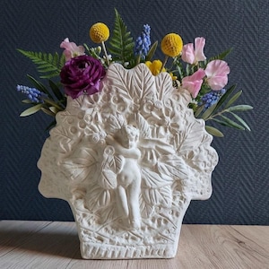 Könnte beinhalten: Eine weiße Keramikvase mit einem Relief eines Putto, umgeben von Blatt- und Blumenmustern. Die Vase ist mit einem farbenfrohen Arrangement aus violetten Ranunkeln, gelben Trommelstöcken, blauen Traubenhyazinthen und rosa Duftwicken gefüllt.