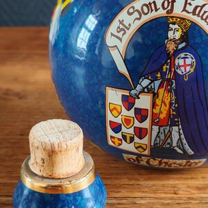 May include: A blue ceramic jar with a cork stopper. The jar features a detailed illustration of a historical figure and heraldic symbols. The stopper has a gold band. The jar is decorated with a mottled blue pattern.