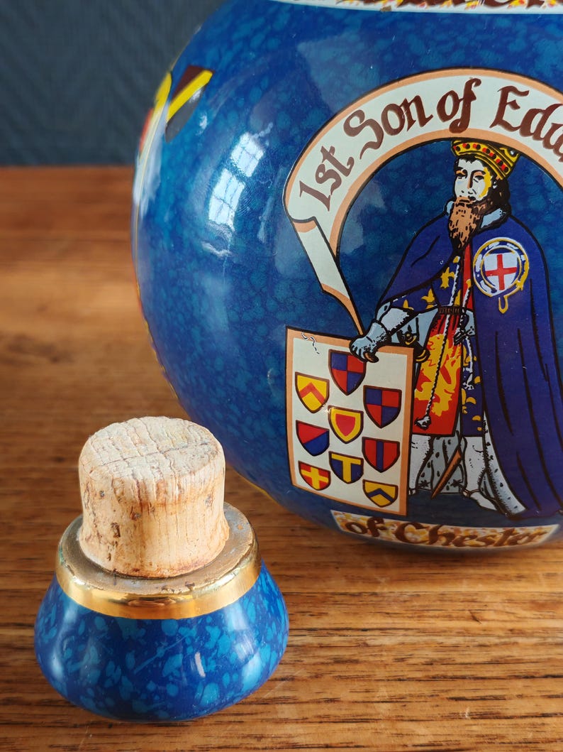May include: A blue ceramic jar with a cork stopper. The jar features a detailed illustration of a historical figure and heraldic symbols. The stopper has a gold band. The jar is decorated with a mottled blue pattern.