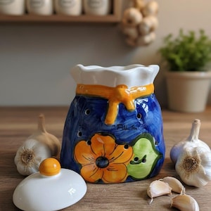 May include: A blue ceramic garlic keeper with a white lid and orange flower design. The keeper has a white ruffled top and a yellow ribbon accent. Several garlic cloves and whole garlic bulbs are arranged around the keeper.