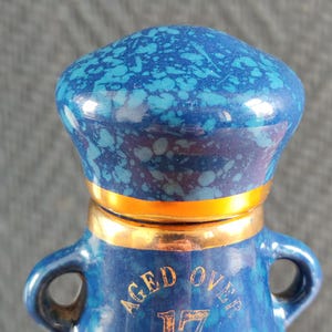 May include: A blue ceramic bottle with a speckled pattern and gold accents. The bottle has a rounded top and two handles. The text "AGED OVER 17 YEARS" is printed on the front.