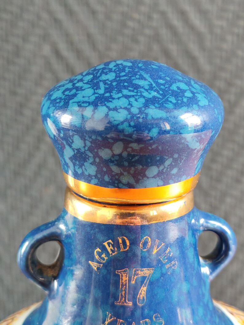 May include: A blue ceramic bottle with a speckled pattern and gold accents. The bottle has a rounded top and two handles. The text "AGED OVER 17 YEARS" is printed on the front.