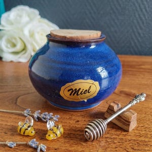 Peut inclure: Un pot à miel en céramique bleu avec un couvercle en liège et une étiquette dorée avec le mot "Miel". Il est accompagné d'un cuillère à miel argentée et de deux petites figurines d'abeilles en verre. Des brins de lavande séchée sont également présents.