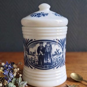 Peut inclure: Un pot en verre opalin blanc avec couvercle, orné d'une impression bleue de style transfert représentant deux personnages en tenue traditionnelle dans un champ de tulipes avec un moulin à vent, et des motifs floraux bleus sur le couvercle.