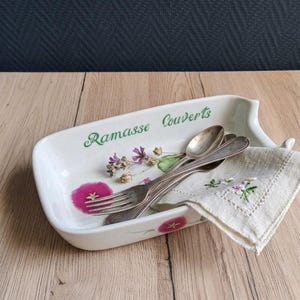 Peut inclure: Un plateau rectangulaire en céramique blanche avec l'inscription verte Ramasse Couverts et des illustrations florales roses et violettes. À l'intérieur se trouvent deux cuillères et une fourchette en métal argenté, ainsi qu'une serviette en lin blanc brodée.