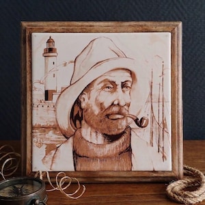 Puede incluir: Un azulejo de cerámica cuadrado enmarcado en madera con una ilustración en tono sepia de un marinero barbudo con sombrero y jersey de cuello alto mientras fuma en pipa. Al fondo, un faro junto al mar con mástiles de barco visibles.